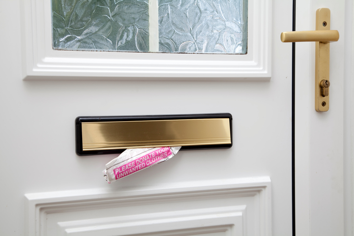 A piece of direct mail sticks partially out of a letter box on a white door. The mail is white with a pink rectangle. Writing in the rectangle in white is, “Please donate your unwanted clothes.”