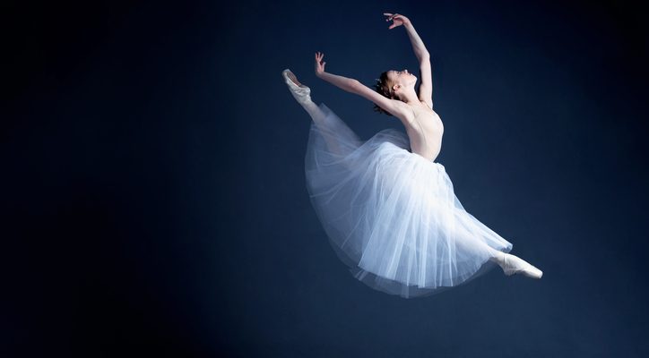 A young ballerina in a beautiful white dress gracefully leaps high through the air in a photo studio.