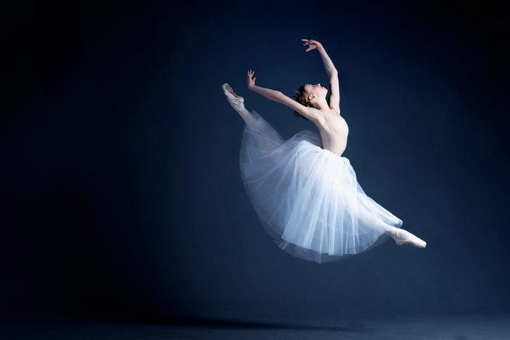 A young ballerina in a beautiful white dress gracefully leaps high through the air in a photo studio.