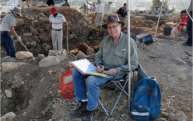 A man at an archeological dig in Israel sits on a chair and smiles at the camera. He has an open folder on his lap and a pen in his hand. People are digging in the background.