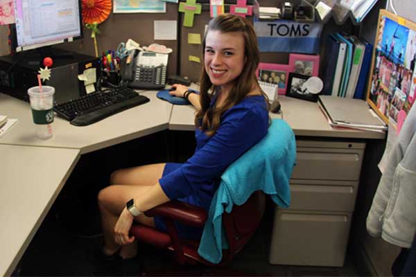 A young woman in a blue blouse sits at her desk in a decorated cubicle and smiles.