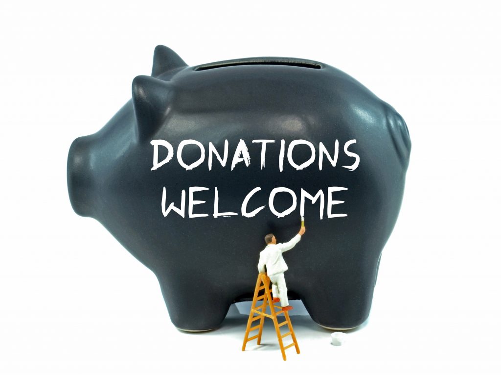 A little man in white stands on a ladder and paints “Donations Welcome” in white on a giant black piggy bank.