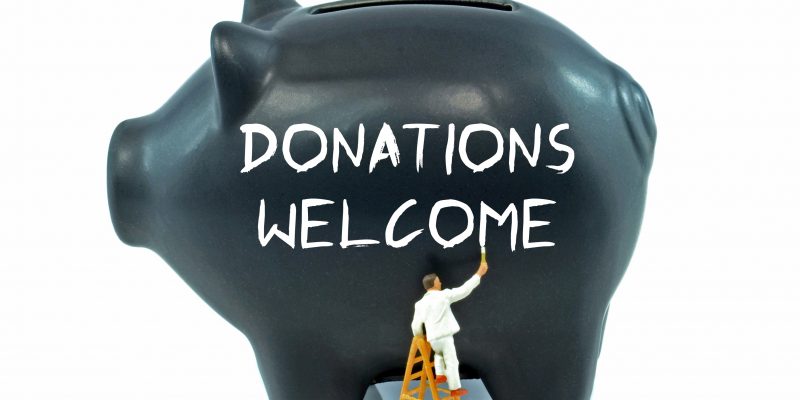 A little man in white stands on a ladder and paints “Donations Welcome” in white on a giant black piggy bank.