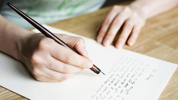 A sheet of paper sits on a desk as a person holds it in place with one hand and begins to write a letter using the quill held in the other. 