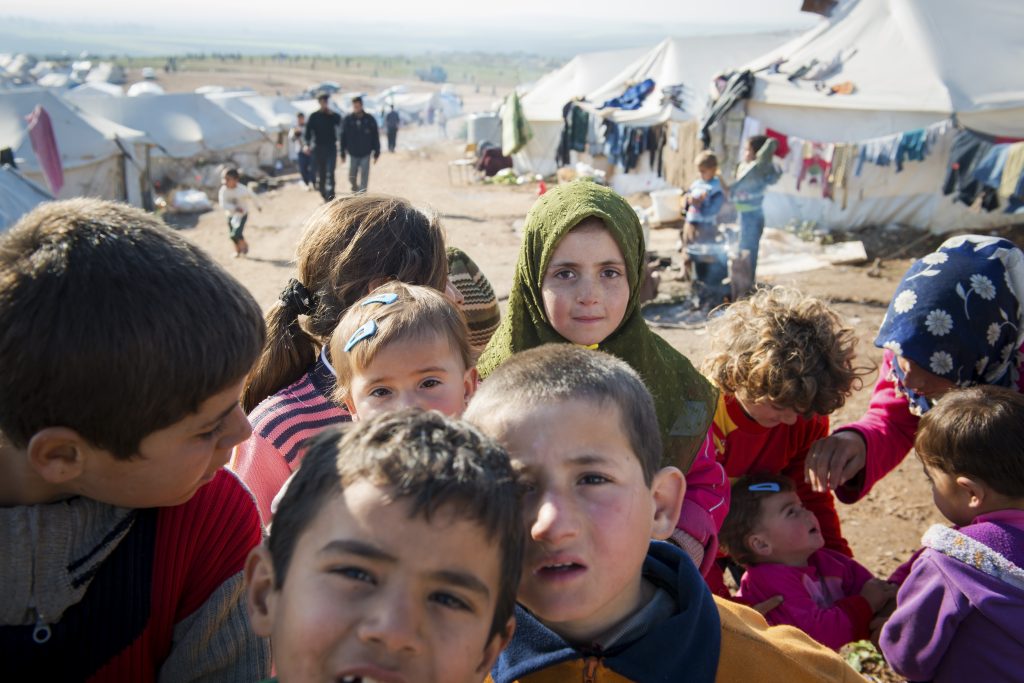 A closeup of about six Syrian refugee children in an internally displaced persons camp. In the background are tents.