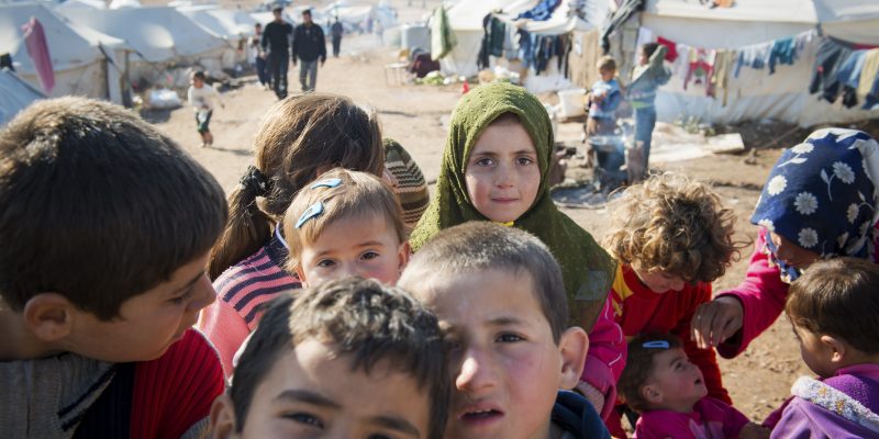 A closeup of about six Syrian refugee children in an internally displaced persons camp. In the background are tents.